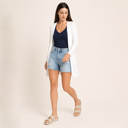 A woman modeling a white open-front cardigan over a navy blue top and light-wash denim shorts, paired with beige slide sandals, against a plain beige studio background.