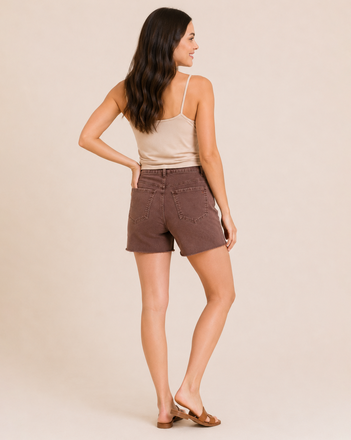 Back view of a woman wearing wine distressed shorts, a beige sleeveless top, and a multicolored plaid shirt on a beige background
