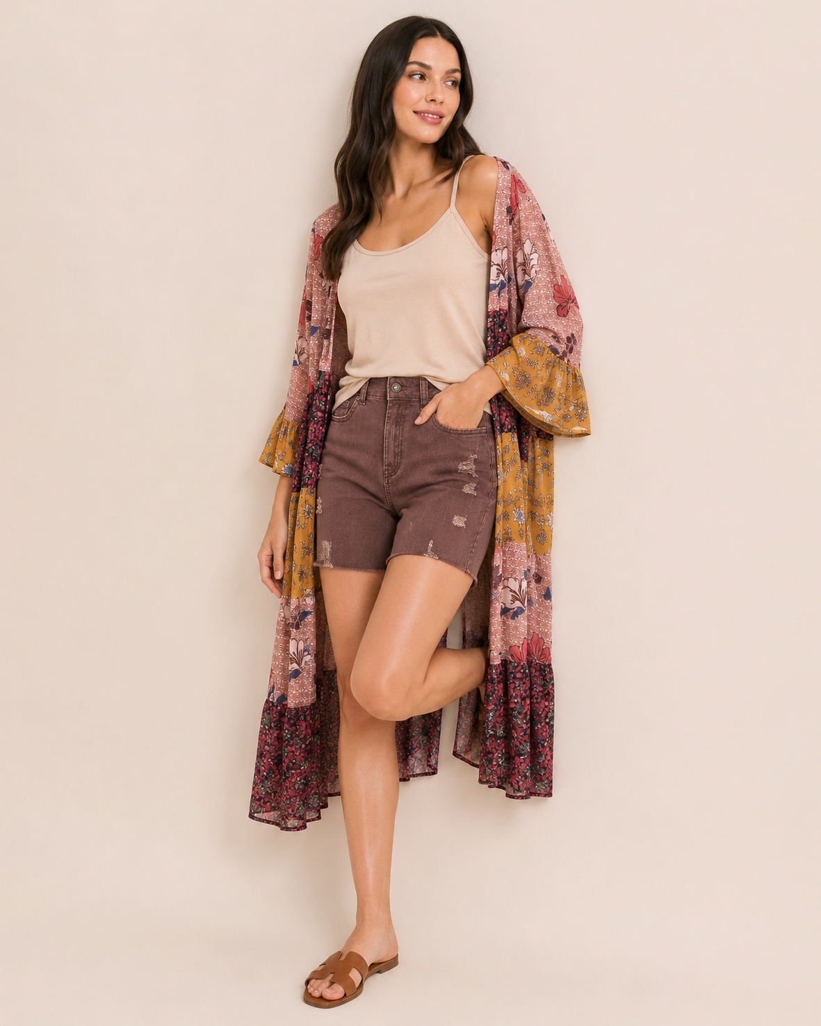 Full-body view of a woman wearing wine distressed shorts, a beige sleeveless top, a multicolored plaid shirt, and brown sandals on a beige background