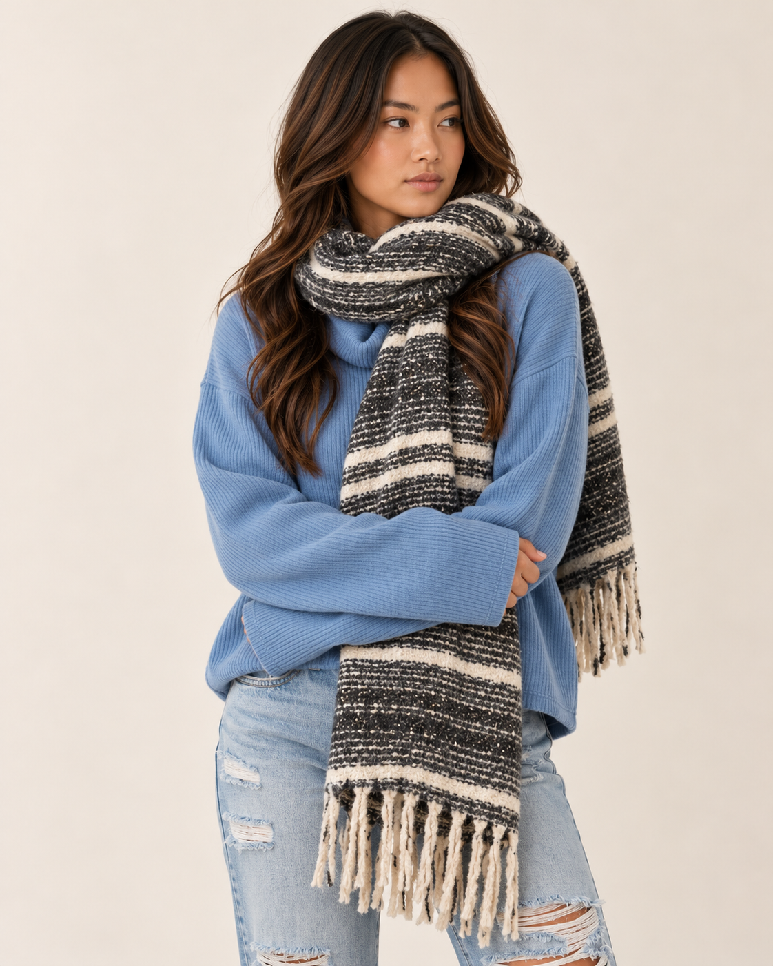 Full-body view of a woman wearing a blue long-sleeve sweater, light-wash distressed jeans, and a black, cream, and gray striped woven scarf with fringe, wrapped around her neck and draped over her shoulders, on a beige background.