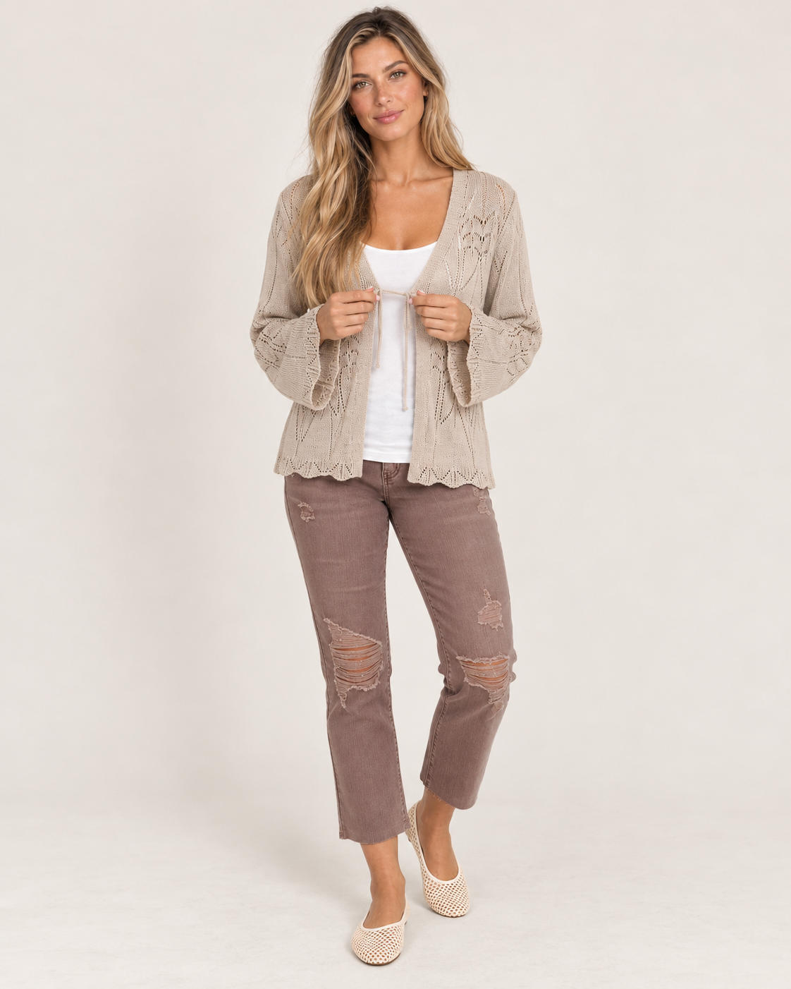 Full-body front view of a woman wearing a taupe open-front knit cardigan with a white top, mauve pants, and beige flats on a beige background