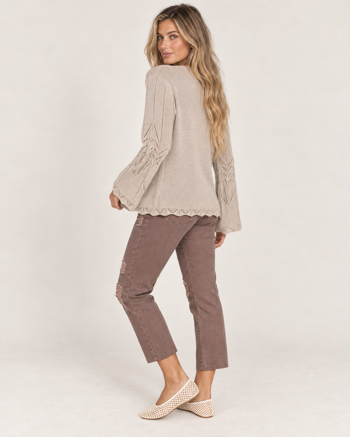 Full-body side view of a woman wearing a taupe open-front knit cardigan with a white top, mauve pants, and beige flats on a beige background