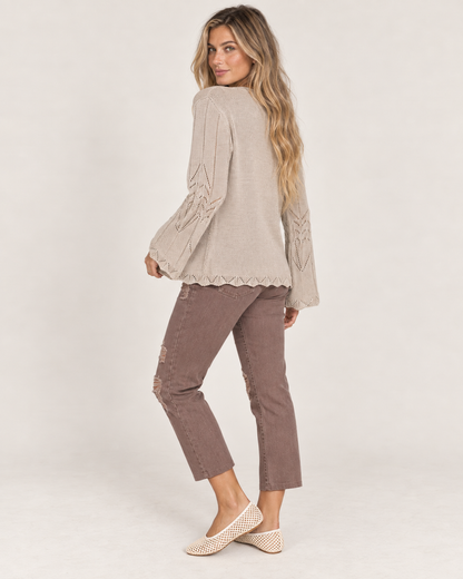 Full-body side view of a woman wearing a taupe open-front knit cardigan with a white top, mauve pants, and beige flats on a beige background