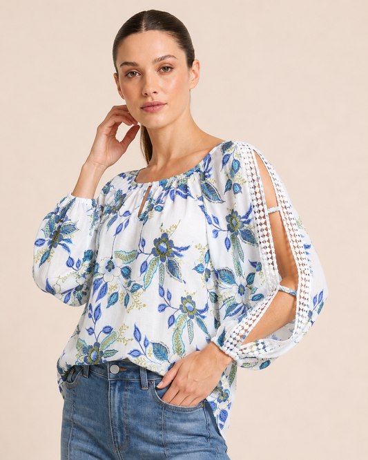 Close-up of a woman wearing a white floral blouse with blue and green flowers, crochet trim sleeve details, and denim jeans on a beige background