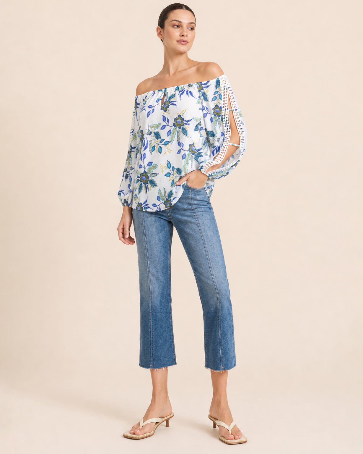 Front view of a woman wearing a white floral blouse with blue and green flowers, crochet trim sleeve details, and blue denim jeans on a beige background