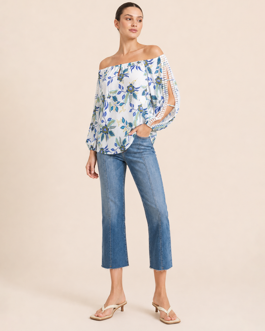 Close-up of a woman wearing a white floral blouse with blue and green flowers, crochet trim sleeve details, and denim jeans on a beige background