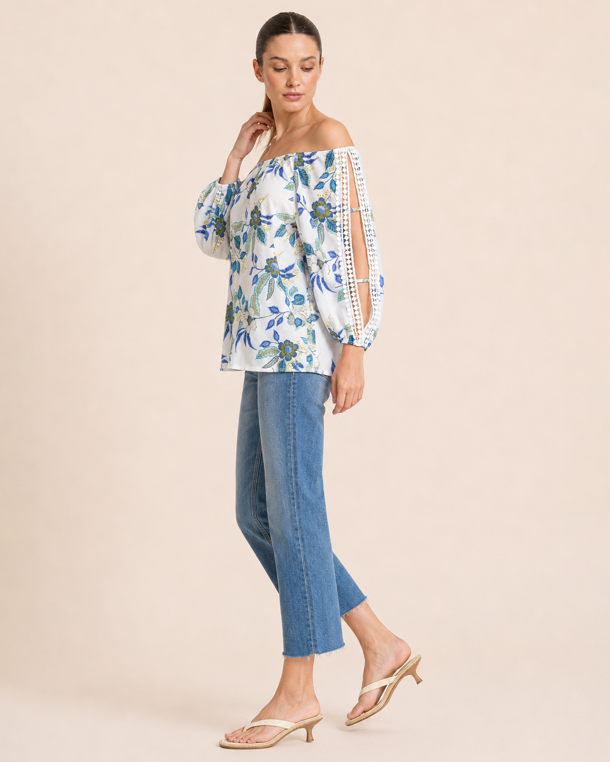 Side view of a woman wearing a white floral blouse with blue and green flowers, crochet trim sleeve details, and blue jeans on a beige background