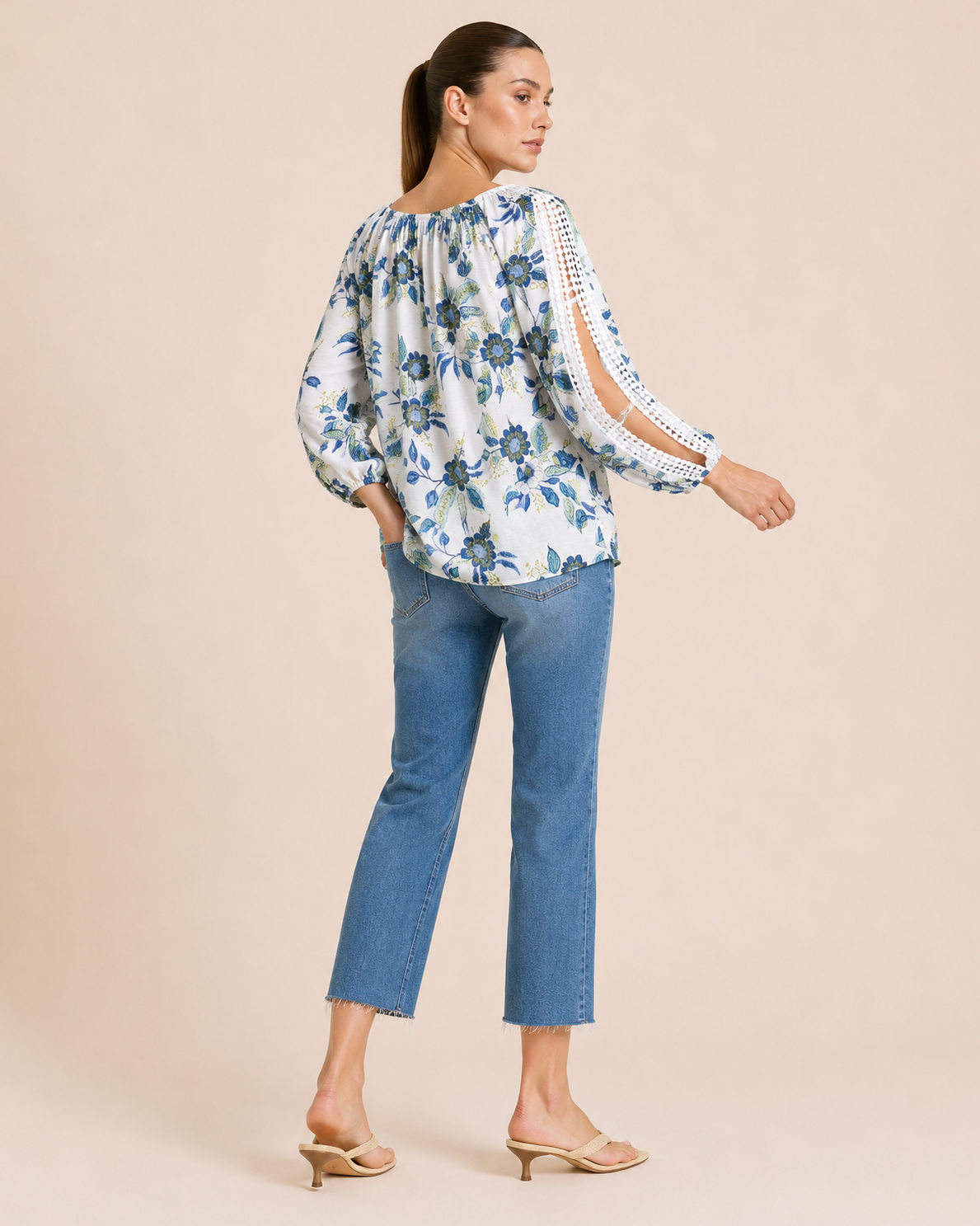 Back view of a woman wearing a white floral blouse with blue and green flowers, crochet trim sleeve details, and blue denim jeans on a beige background