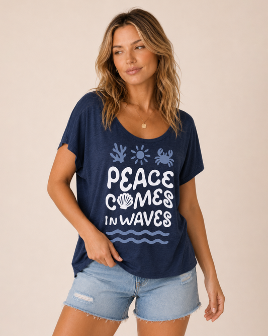 Front view of a woman wearing a navy graphic tee featuring a "Peace Comes in Waves" design and light wash denim shorts on a beige background