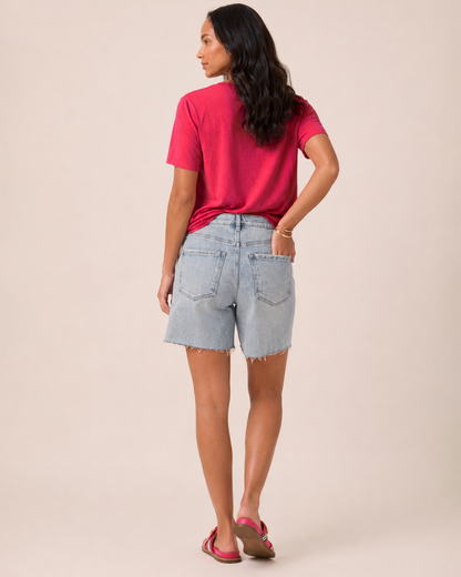 Side view of a woman wearing light wash denim shorts, a coral short-sleeve blouse, and brown sandals on a beige background