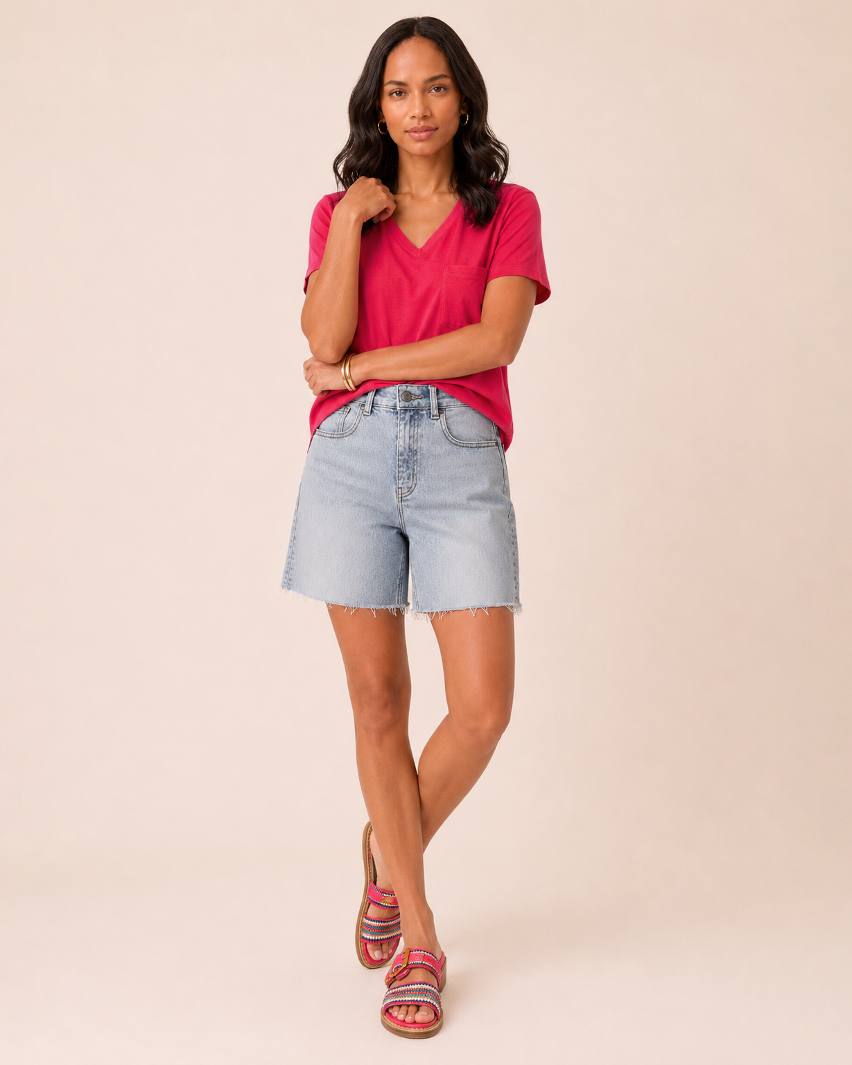 Full-body view of a woman wearing light wash denim shorts, a coral short-sleeve blouse, and brown sandals on a beige background