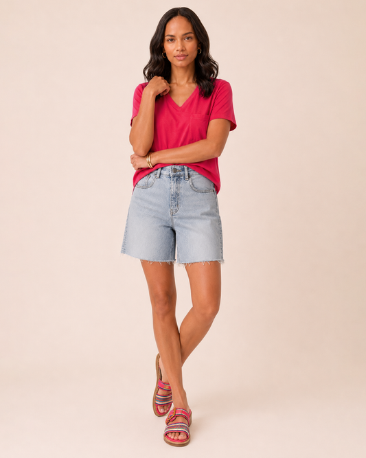 Full-body view of a woman wearing light wash denim shorts, a coral short-sleeve blouse, and brown sandals on a beige background