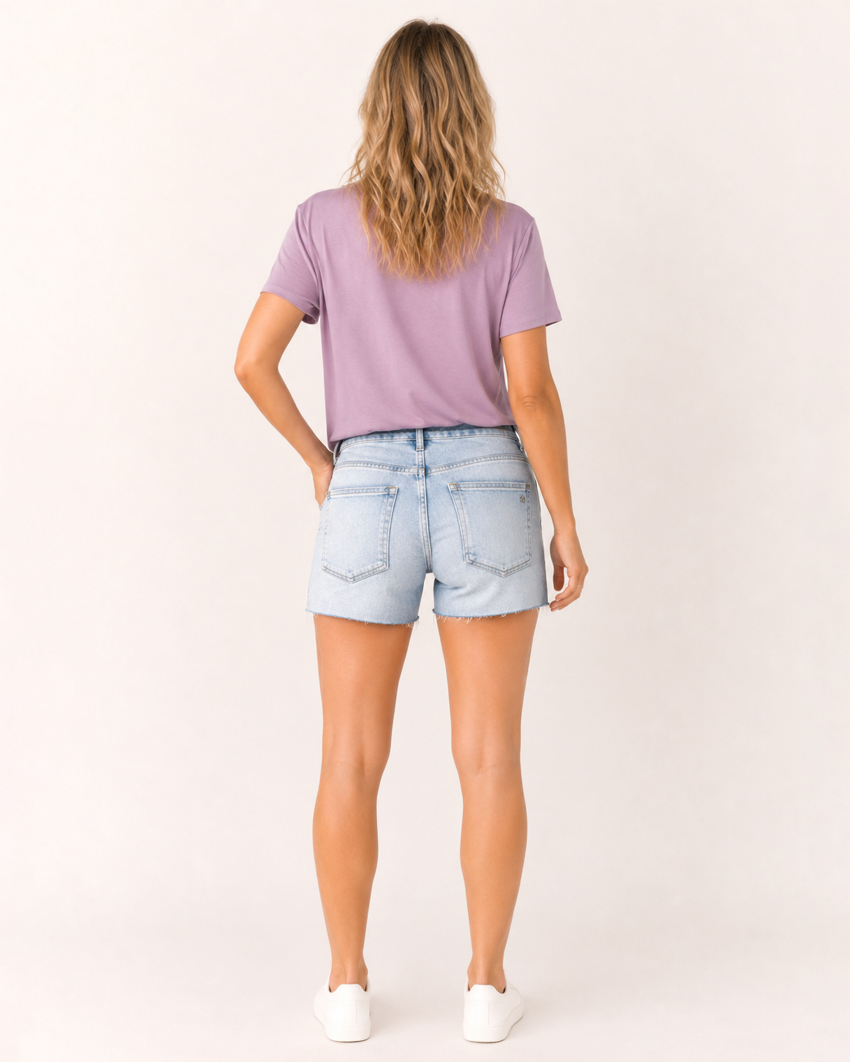 Back view of a woman wearing light wash distressed denim shorts and a lavender V-neck t-shirt on a beige background