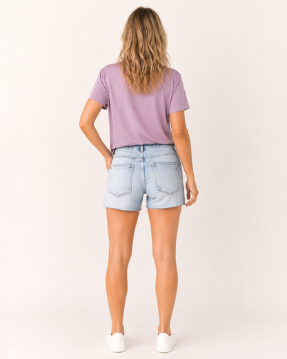 Back view of a woman wearing light wash distressed denim shorts and a lavender V-neck t-shirt on a beige background