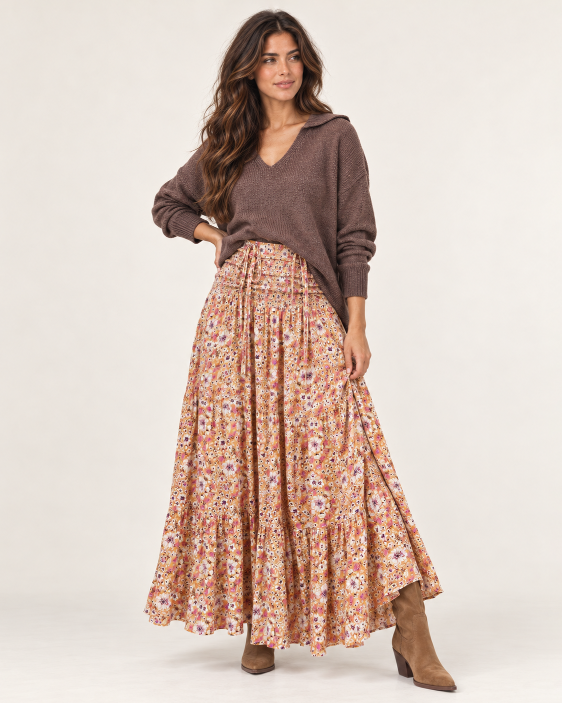 Full-body side view of a woman wearing a mauve V-neck knit cardigan with a floral pleated midi skirt and tan flat shoes on a beige background.