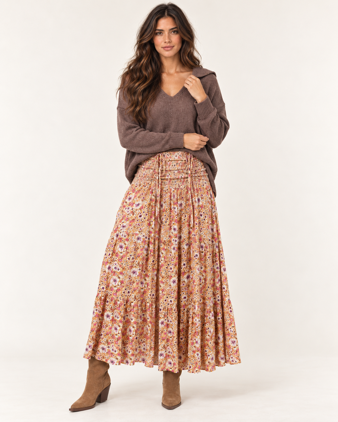 Full-body front view of a woman wearing a mauve V-neck knit cardigan with a floral pleated midi skirt and tan flat shoes on a beige background.