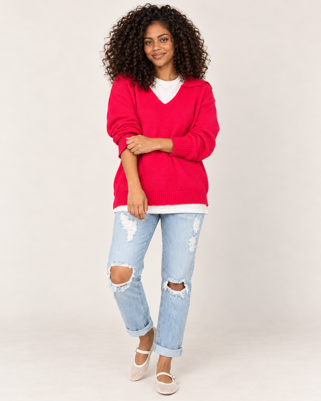 Full-body front view of a woman wearing a bright red long-sleeve pullover with light-wash distressed jeans and dark sneakers on a beige background.