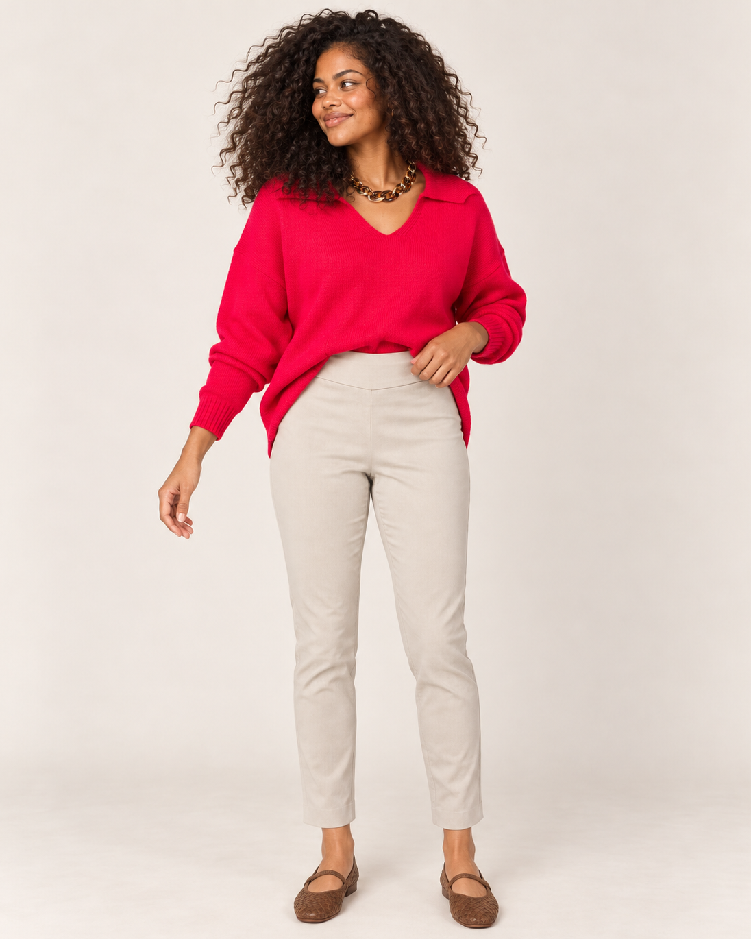 Full-body front view of a woman wearing a bright red long-sleeve pullover with beige straight-leg pants and tan flat shoes on a beige background.