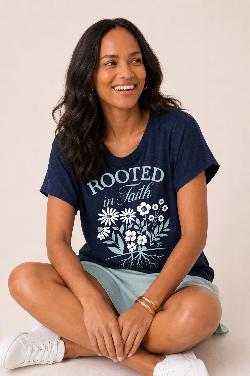 Close-up of a woman wearing a navy blue graphic tee featuring a "Rooted in Faith" floral design and a light sage skirt on a beige background