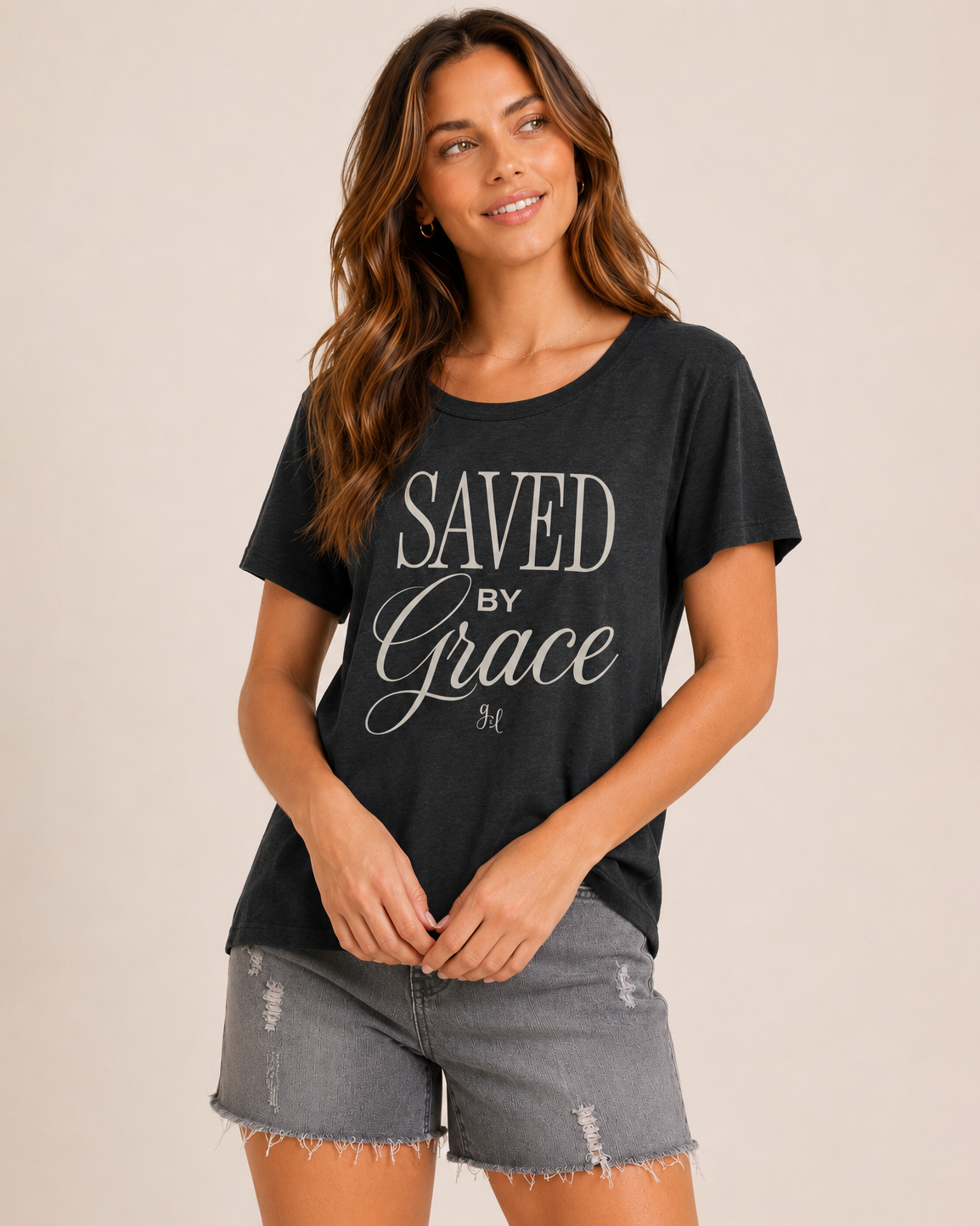 Close-up of a woman wearing a charcoal graphic tee featuring a "Saved By Grace" inspirational design and grey distressed denim shorts on a beige background
