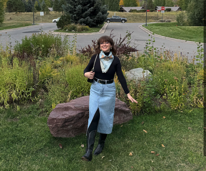 Woman standing outdoors in a garden with a road and buildings in the background