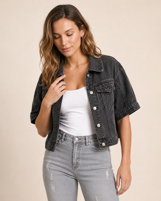 Close-up of a woman wearing a black short sleeve denim jacket over a white tank top and light grey distressed jeans on a beige background