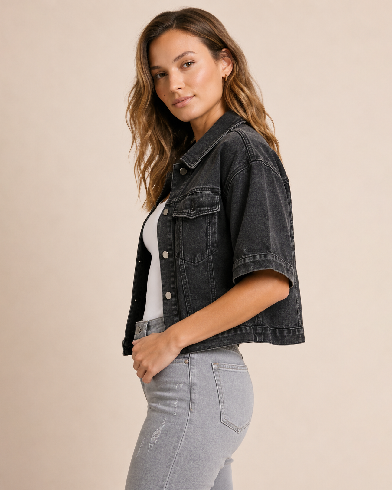 Side view of a woman wearing a black short sleeve denim jacket over a white tank top and light grey distressed jeans on a beige background