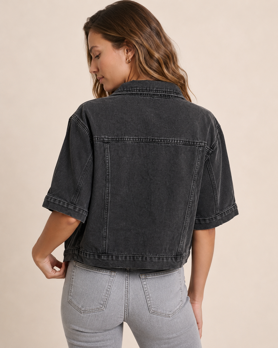 Back view of a woman wearing a black short sleeve denim jacket and light grey distressed jeans on a beige background