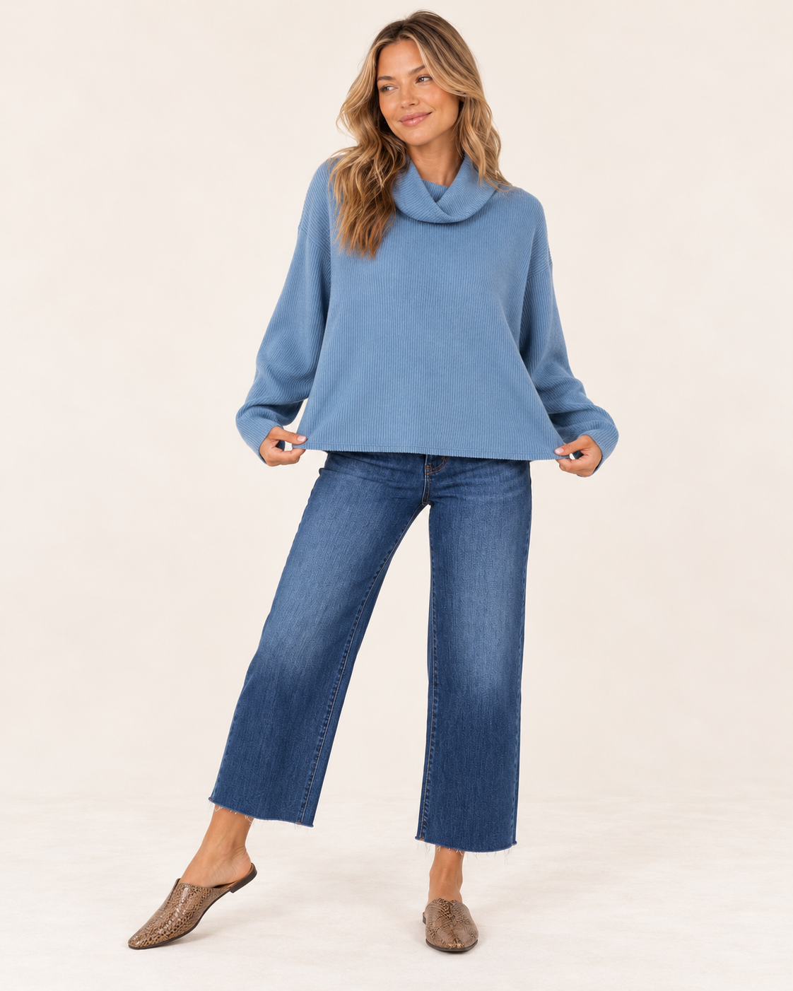 A woman modeling a light blue cowl-neck knit sweater and blue wide-leg cropped jeans with taupe mule flats, standing against a plain off-white studio background.