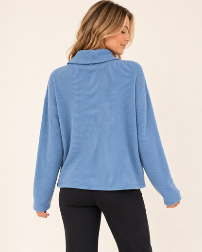 A woman shown from the back wearing a light blue ribbed knit mock neck sweater and black pants, set against a plain cream background.