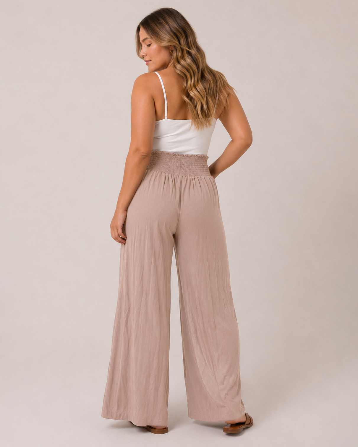 A woman wearing high-waisted, wide-leg dusty pink pants and a white spaghetti-strap tank top, shown from the back against a plain beige studio background.