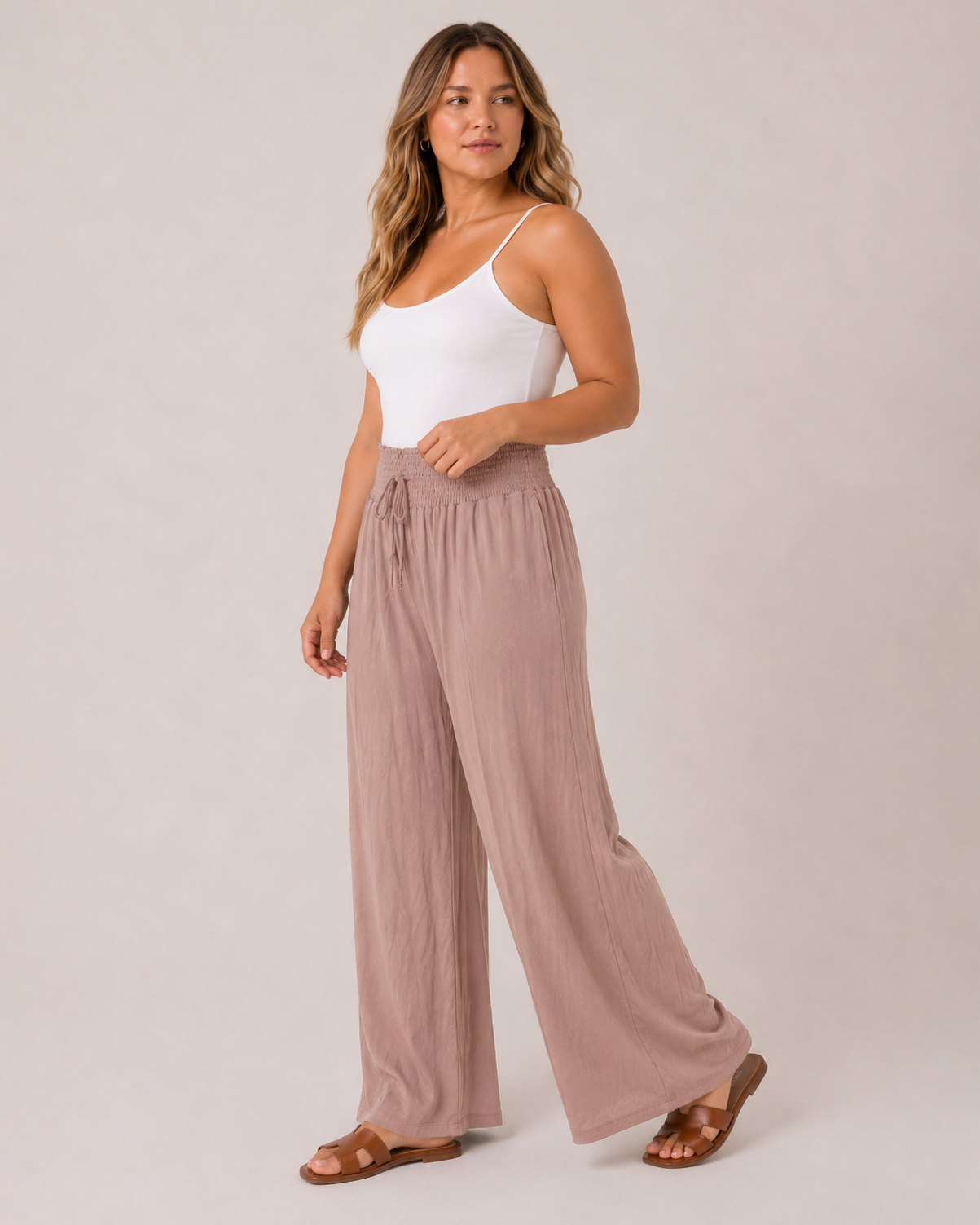 A woman modeling a pair of high-waisted, wide-leg pleated pants in a dusty rose color, paired with a white camisole and brown sandals, against a solid light beige studio background.