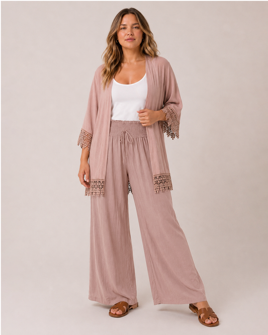 A woman modeling a dusty rose two-piece lounge set consisting of a lightweight open-front cardigan with lace trim and matching wide-leg drawstring pants, styled over a white tank top and brown slide sandals against a solid light beige studio background.