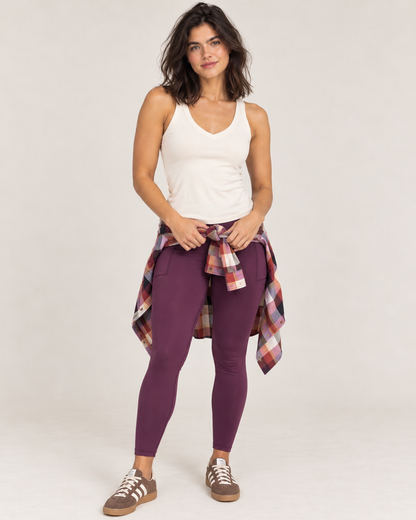 A woman modeling purple leggings with a white tank top and a red plaid shirt tied around her waist, standing against a light gray studio background.
