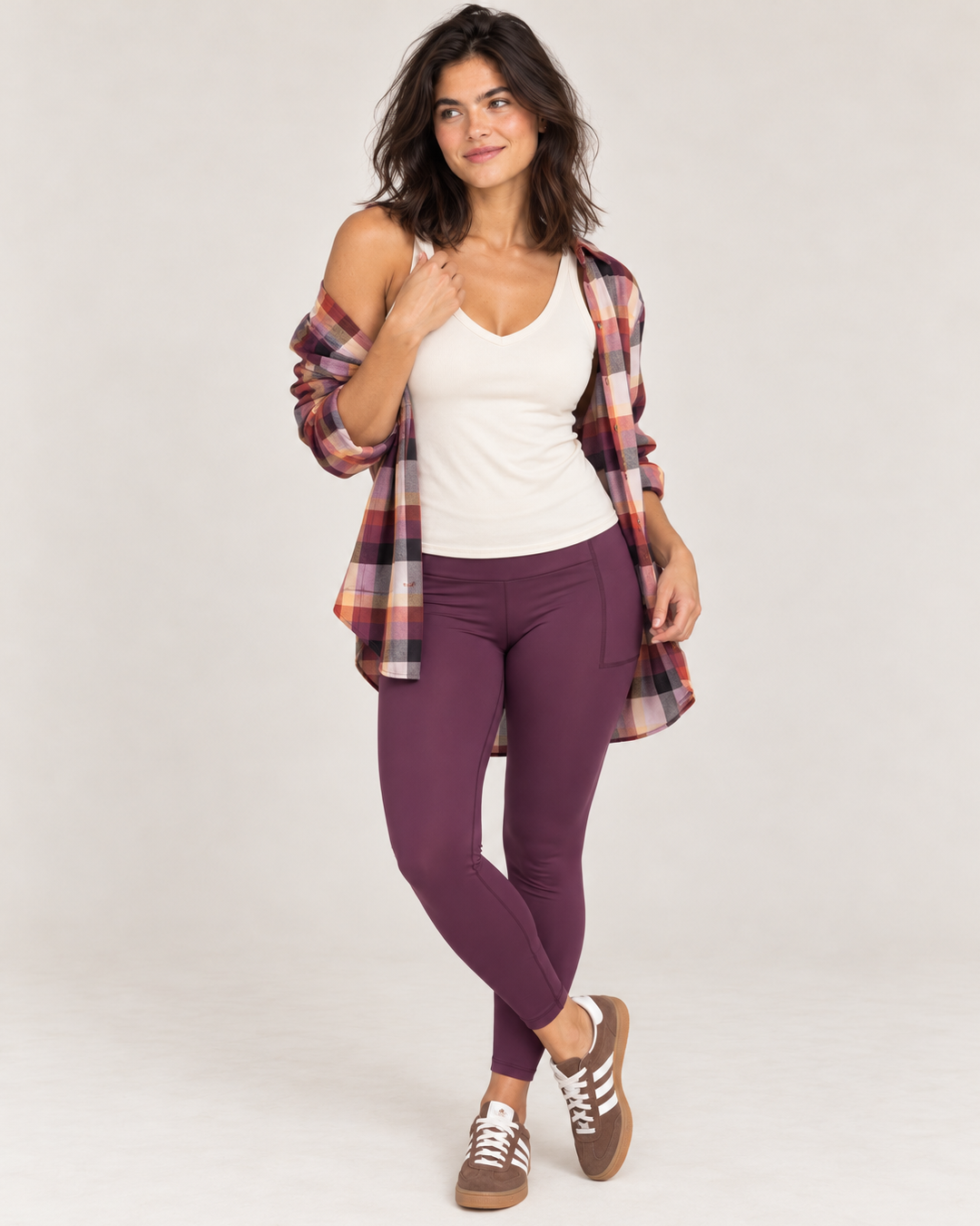 A woman modeling a casual outfit consisting of a white tank top, purple leggings, a multicolor plaid flannel shirt, and brown sneakers, photographed against a light gray studio background.