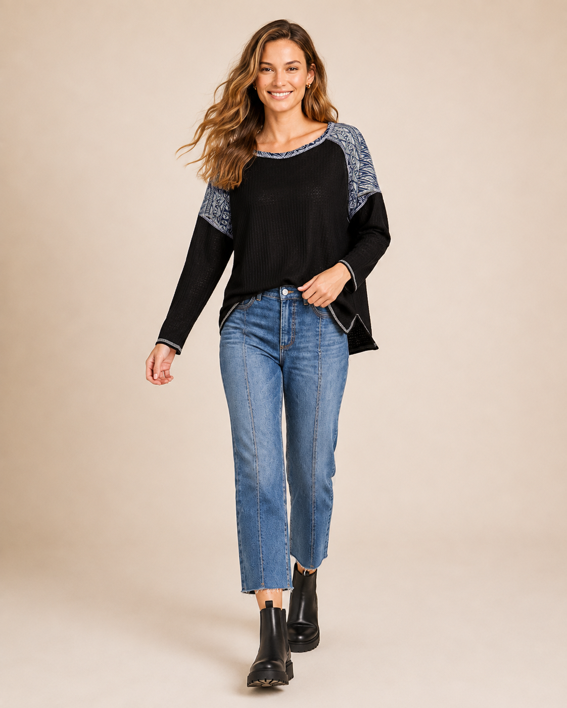 Full-body view of a woman wearing a black and gray color-block knit top with blue jeans and black ankle boots on a beige background
