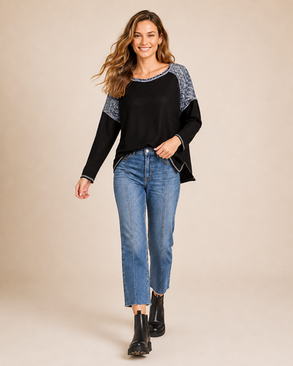Full-body view of a woman wearing a black and gray color-block knit top with blue jeans and black ankle boots on a beige background