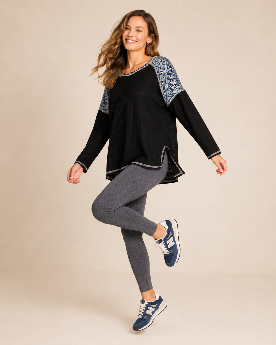 Full-body view of a woman wearing a black and gray color-block knit top with gray leggings and navy and white sneakers on a beige background