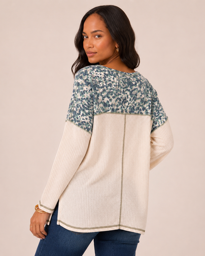 Back view of a woman wearing a white knit top with blue and gray patterned sleeves and blue jeans on a beige background