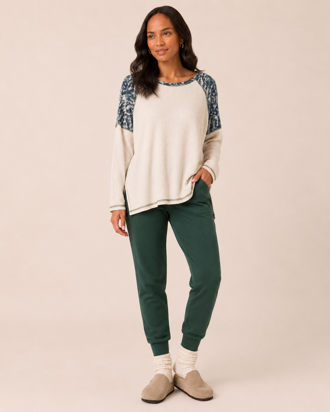 Full-body front view of a woman wearing a cream knit pullover with blue floral-patterned sleeves, dark green jogger pants, cream socks, and taupe slip-on shoes on a beige background