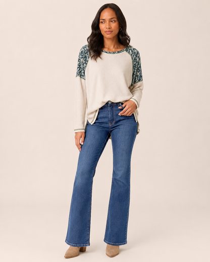 Full-body front view of a woman wearing a white knit top with blue and gray patterned sleeves, blue flared jeans, and tan heeled shoes on a beige background