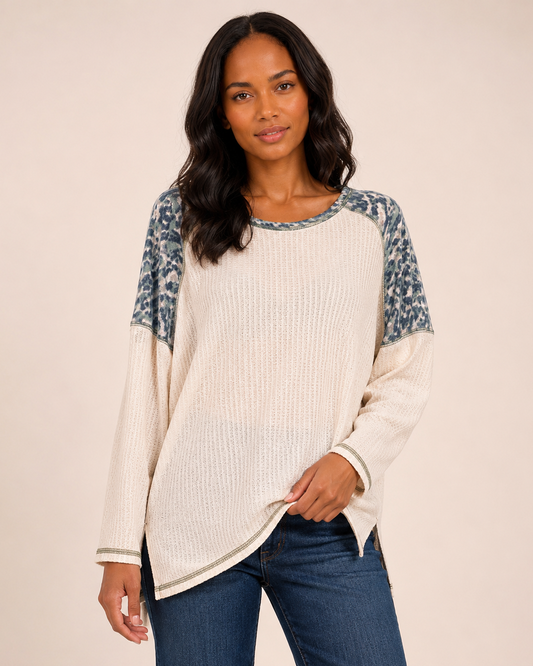 Front view of a woman wearing a white knit top with blue and gray patterned sleeves and blue jeans on a beige background