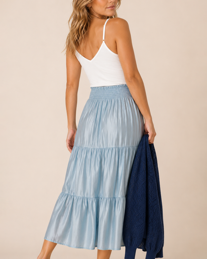 A woman wearing a light blue tiered midi skirt and a white camisole, holding a navy blue sweater against a plain beige studio background.