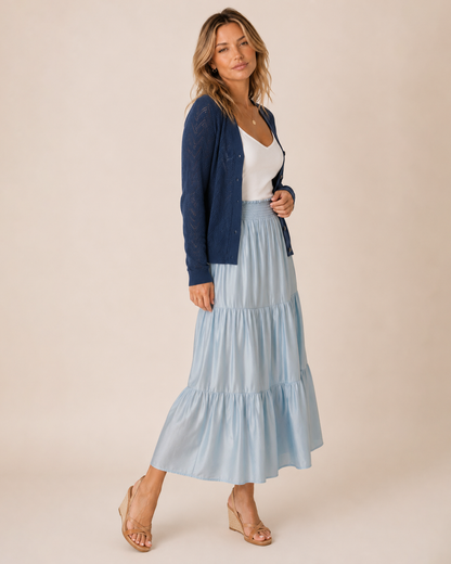 A woman models a light blue tiered maxi skirt paired with a navy blue cardigan, white top, and beige wedge sandals against a solid beige studio background.