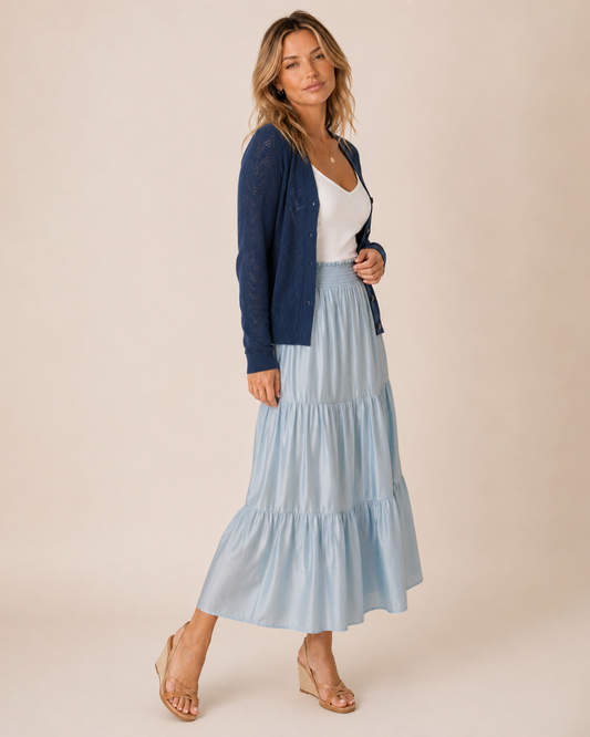 A woman models a light blue tiered maxi skirt paired with a navy blue cardigan, white top, and beige wedge sandals against a solid beige studio background.