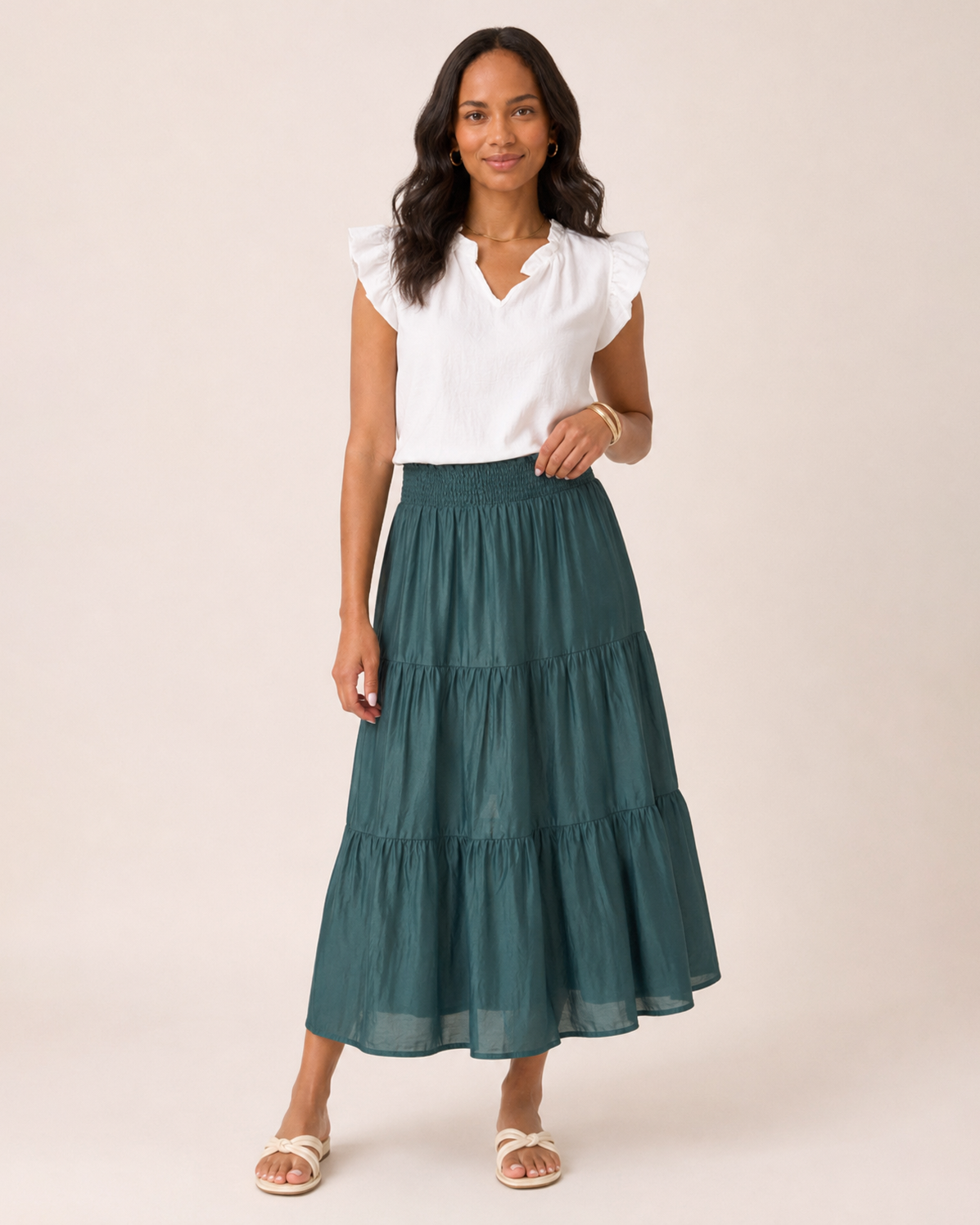 Full-body view of a woman wearing an ivy tiered maxi skirt, a white flutter sleeve blouse, and tan sandals on a beige background