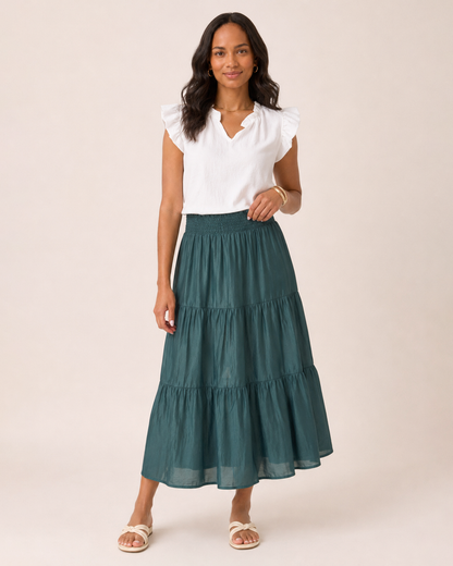 Full-body view of a woman wearing an ivy tiered maxi skirt, a white flutter sleeve blouse, and tan sandals on a beige background