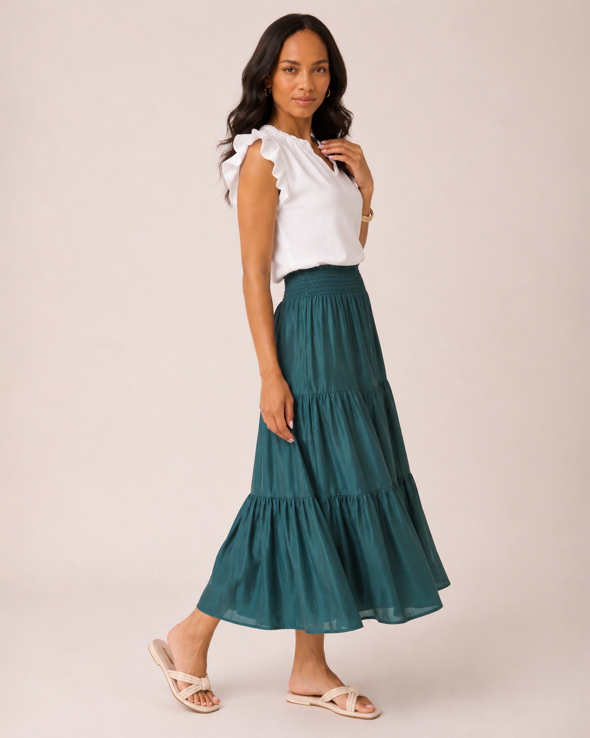 Angled full-body view of a woman wearing an ivy tiered maxi skirt, a white flutter sleeve blouse, and tan sandals on a beige background