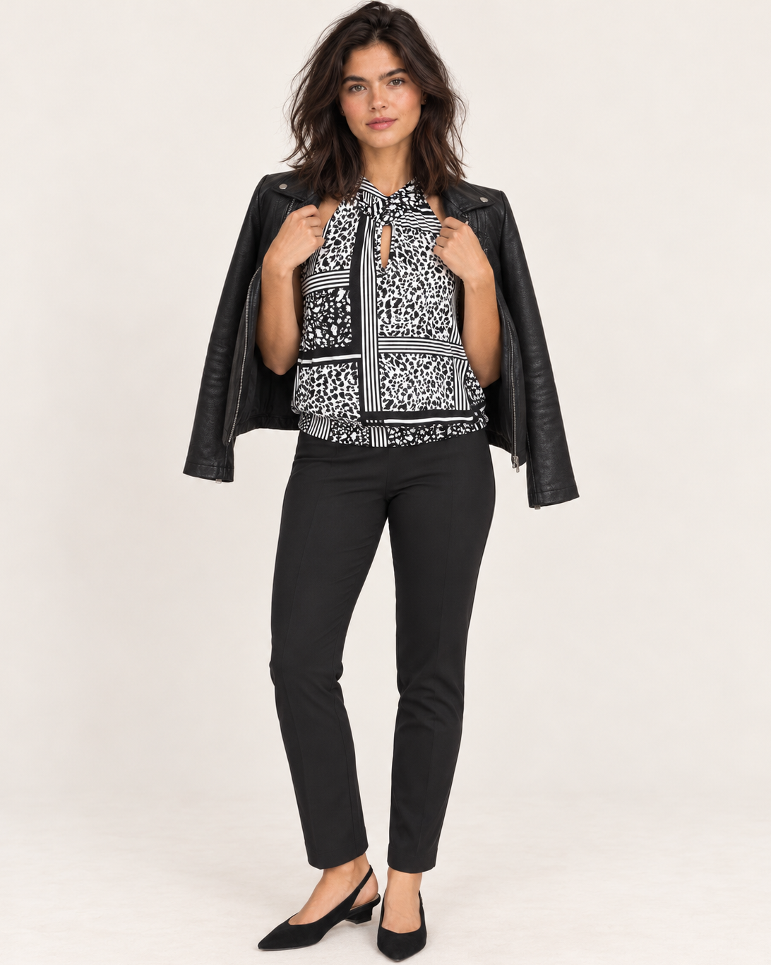 A woman modeling a black leather jacket worn open over a black and white patterned blouse, paired with black pants and black heels, against a plain light background.