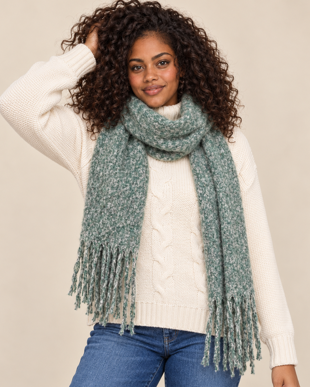 Cropped front view of a woman wearing a sage green marled knit scarf with long fringed ends wrapped around her neck, paired with a cream cable-knit sweater and blue jeans on a beige background.